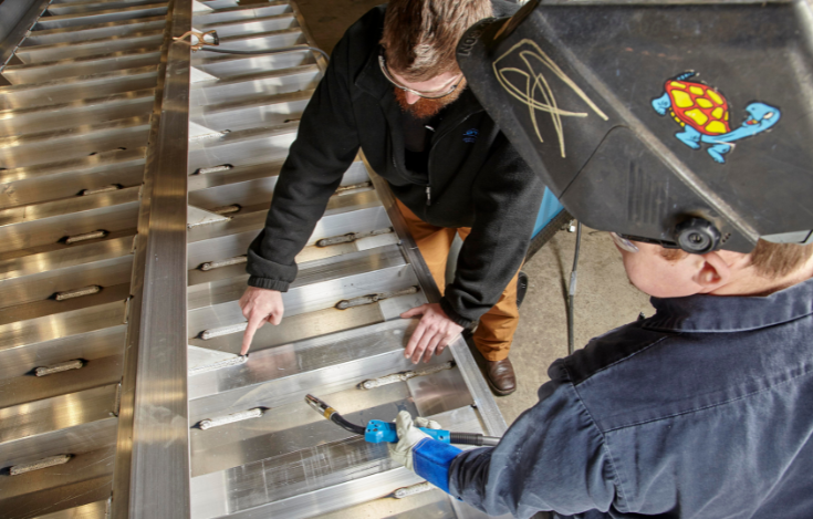 Two welders using OTC DAIHEN Welbee welding equipment power source examine weld quality on aluminum.